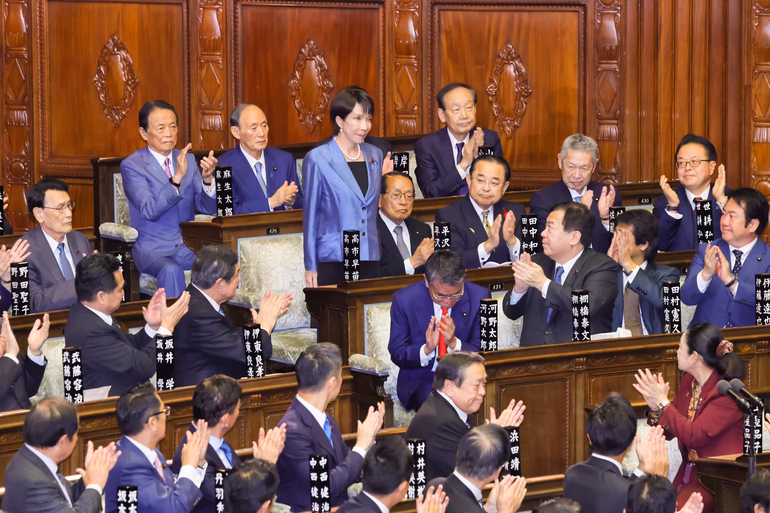 [高市総理誕生の瞬間の衆議院の様子。起立した高市氏に拍手が送られている。]