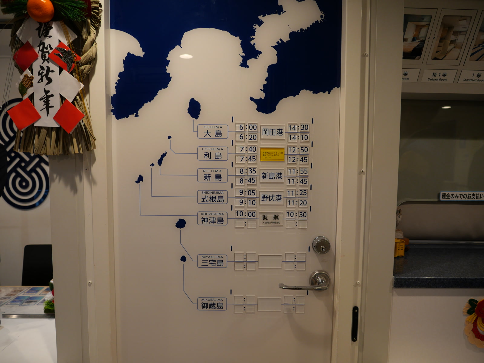 船内フロントの白い扉に掲示された時刻表。各島における発着予定時刻が記載されているが、利島は「天候状況によりましては運行できない場合がございます」と書かれている。