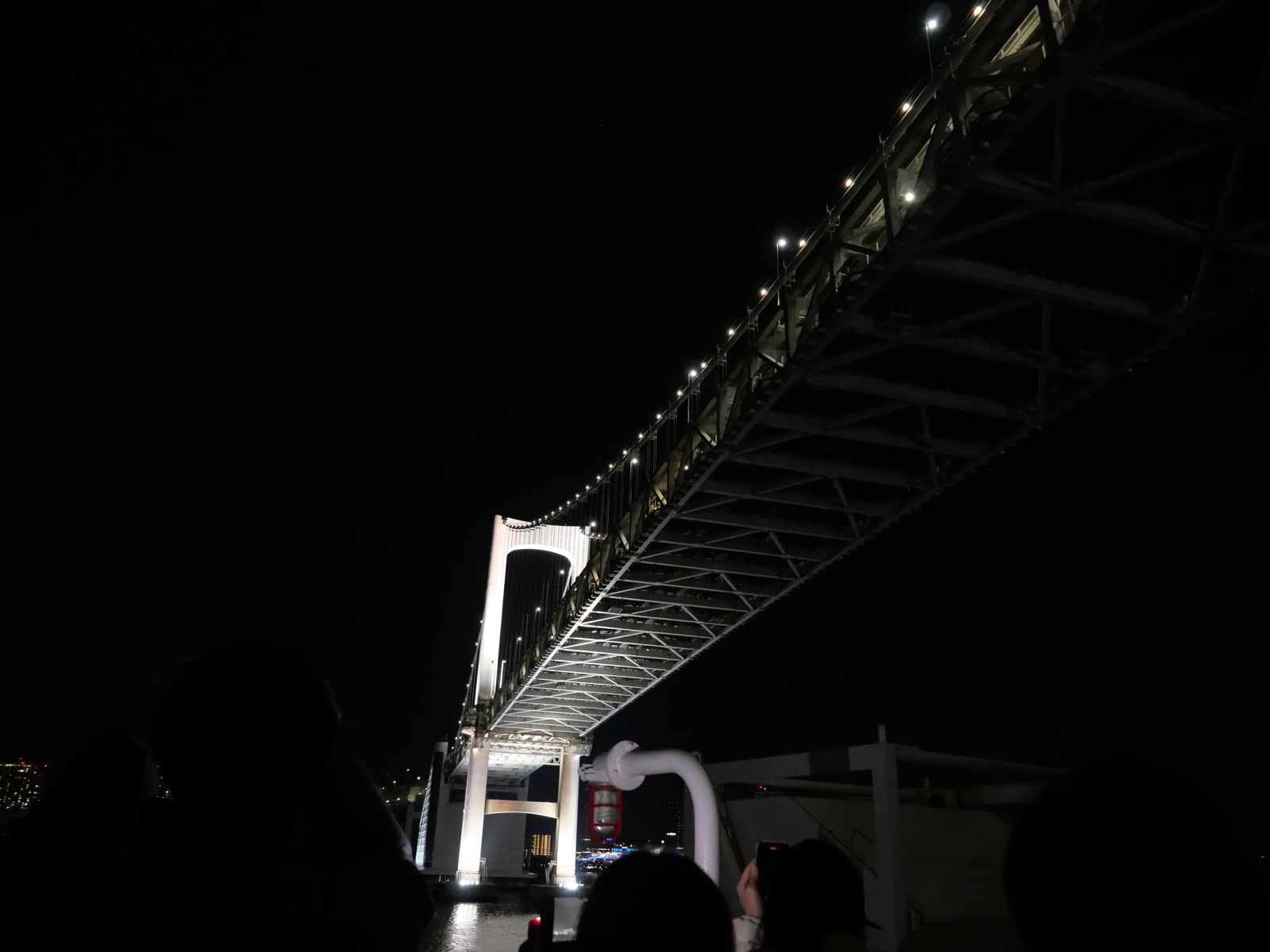 夜間に船の甲板から見上げたレインボーブリッジ。漆黒の夜空を背景に、ライトアップされた橋の白い構造と点々と続く照明が、巨大なアーチを描いて頭上に架かっている。