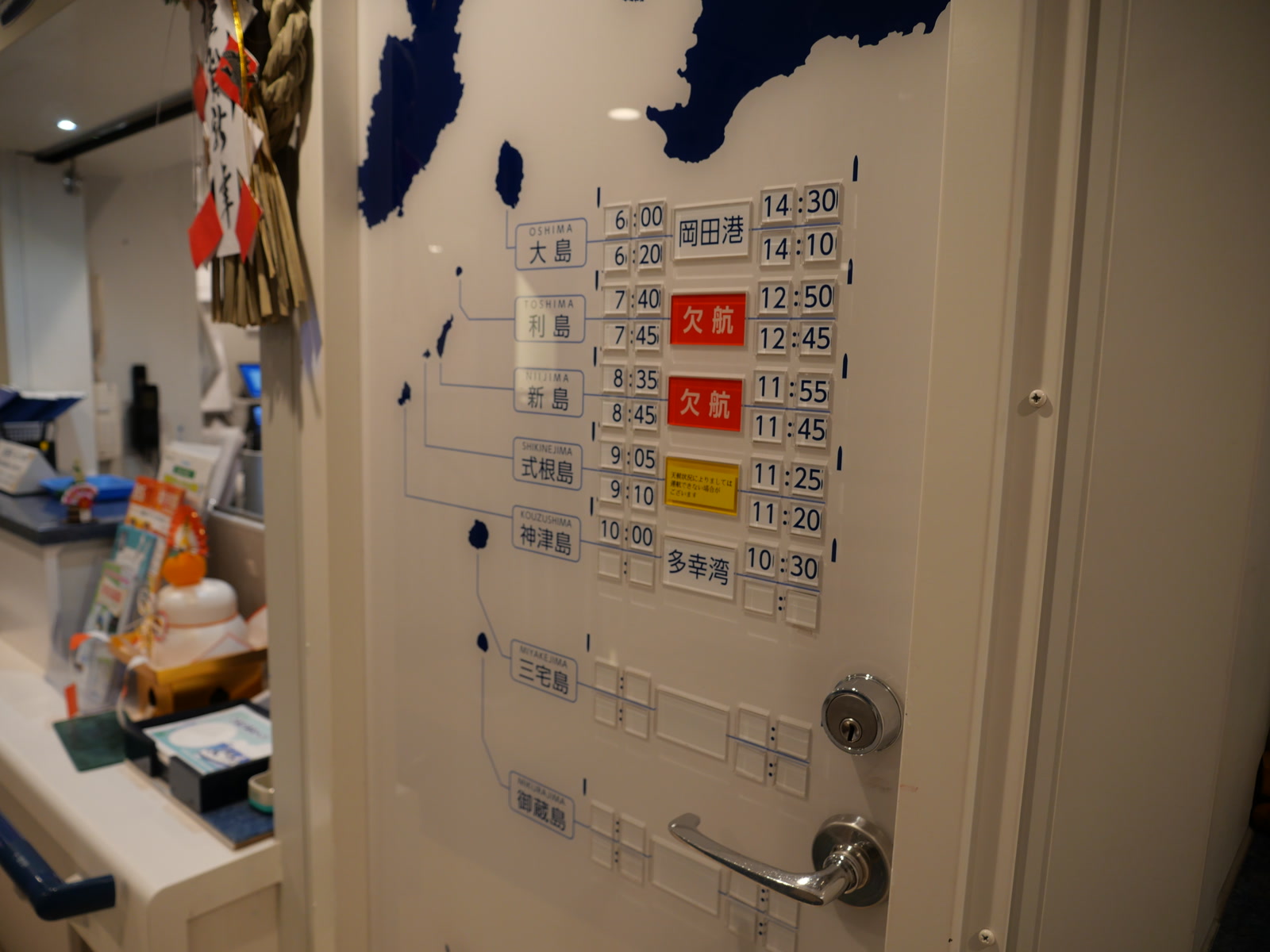 船内フロントの白い扉に掲示された時刻表。利島、新島が「欠航」表記になり、式根島も「天候状況によりましては運行できない場合がございます」表記に変化している。